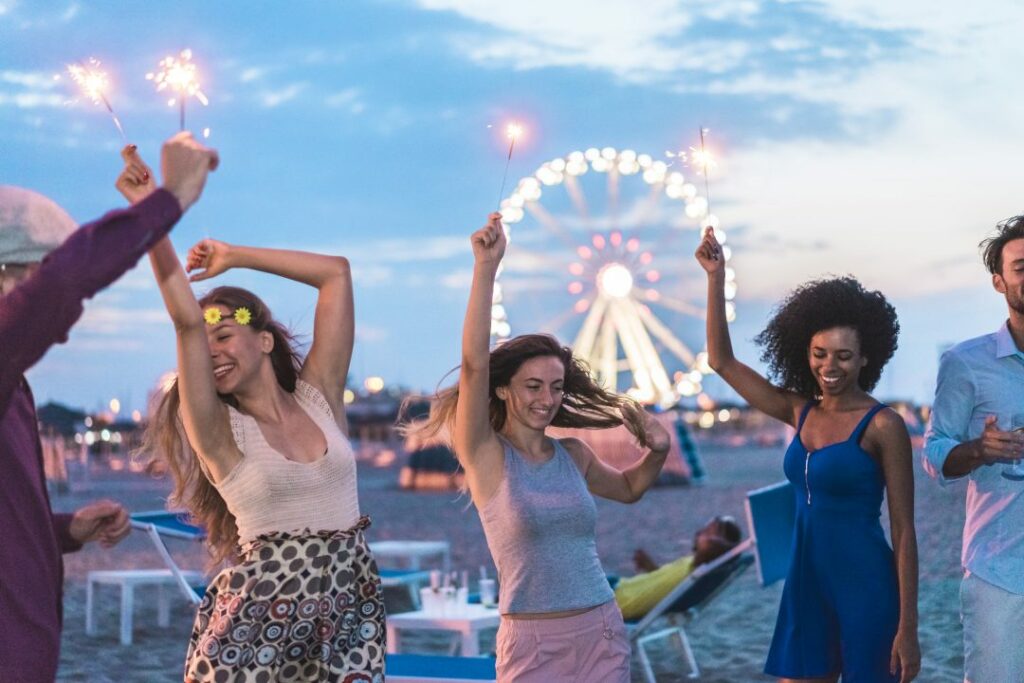 Happy friends making beach party after sunset with fireworks and drinking champagne - Young people having fun dancing outdoor - Soft focus on center girl face - Youth and festival concept